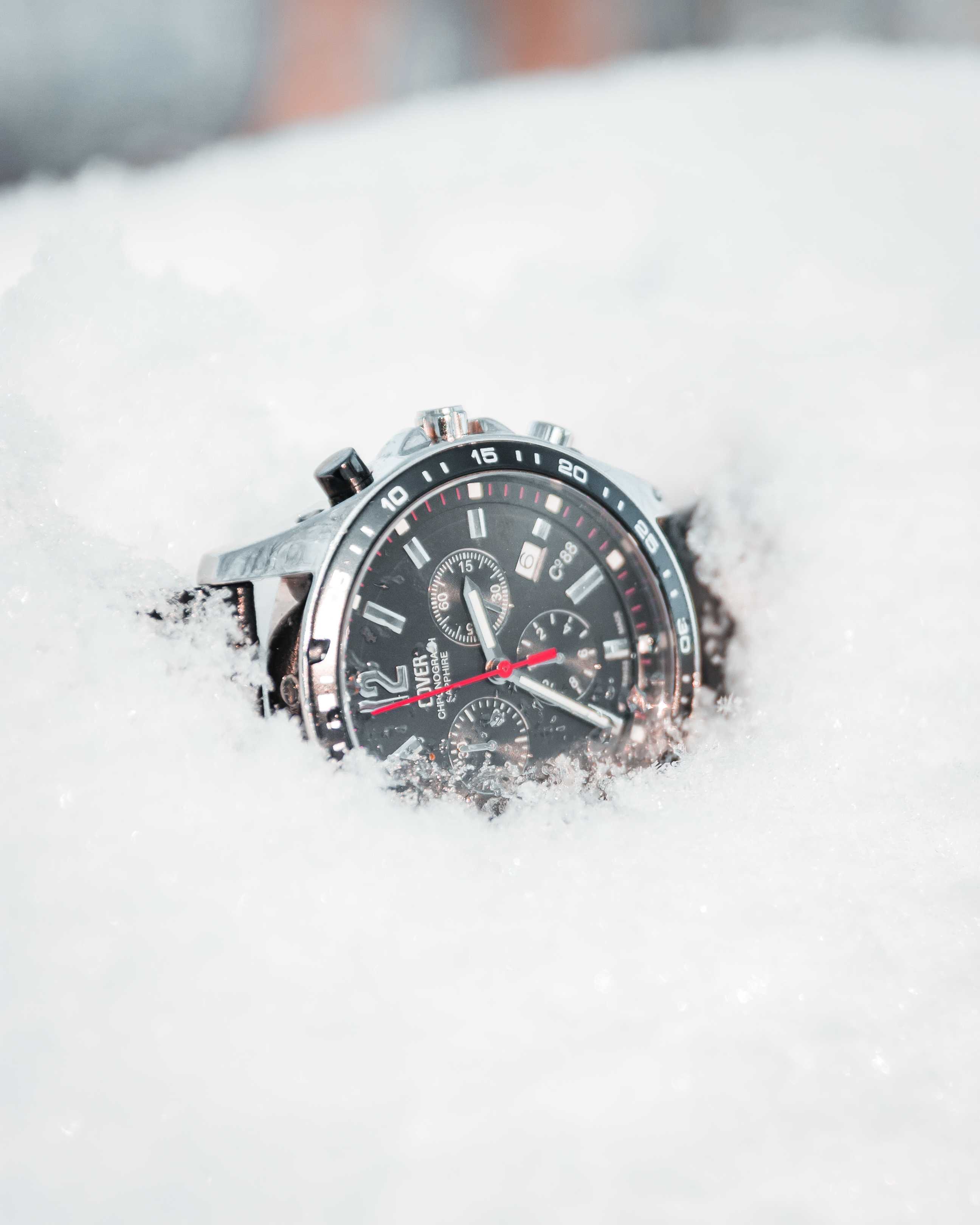 A black and silver watch sitting in the snow. Watch has a single red hand and two white hands.