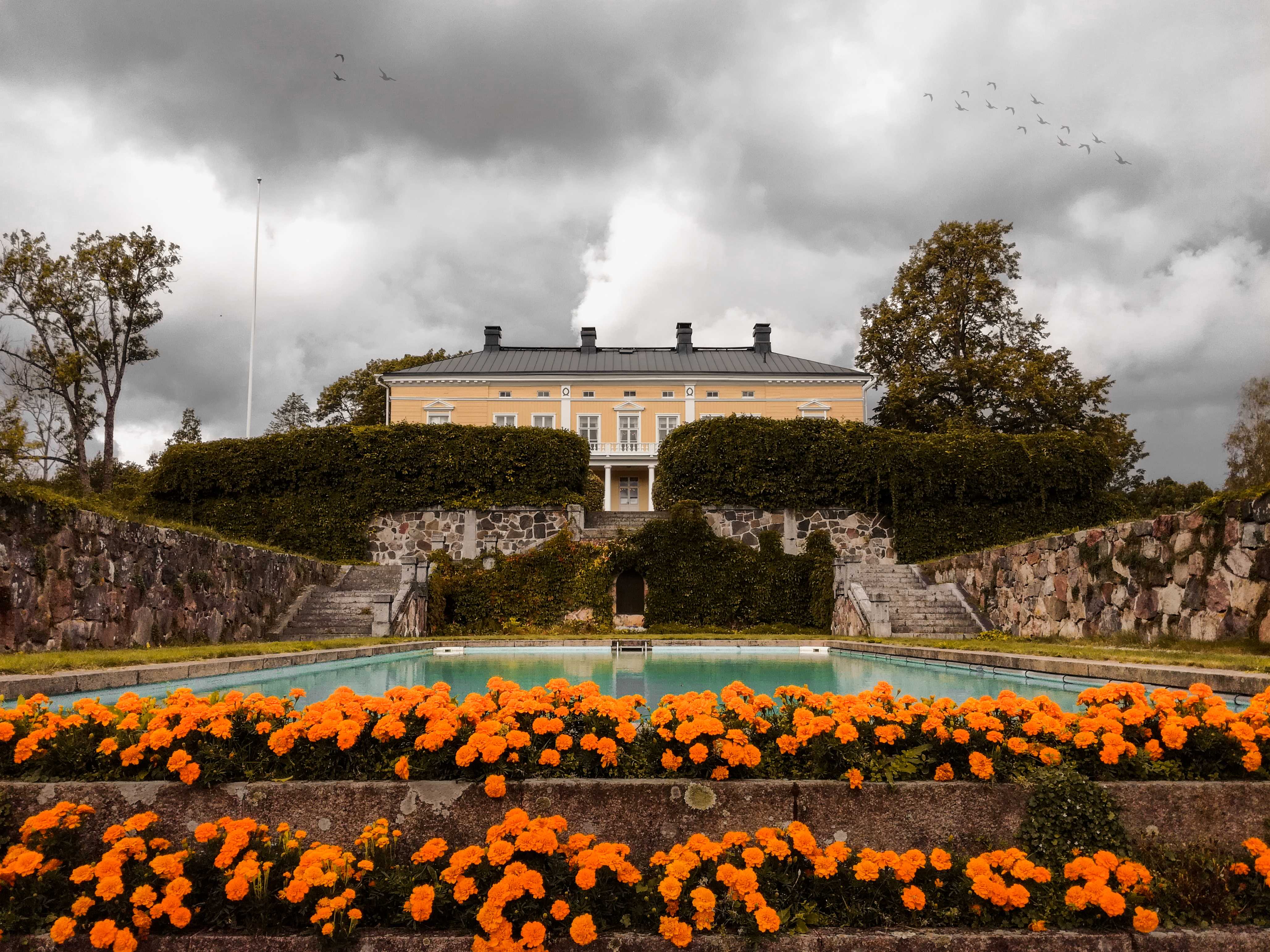 Photograph of the Kirkniemi manor. On the bottom there are orange flowers in two steps leading up to a pool. On either side of the pool there is a stone paved paths with stone walls on outer side of the path. At the end of the pool and the path there are stone steps leading up to the manor. From halfway up the image there can be seen hdges in front of the manor with gray skies. Few bird are flying up in the air. Photo is shoot in an upwards angle so that it rises towards the manor.