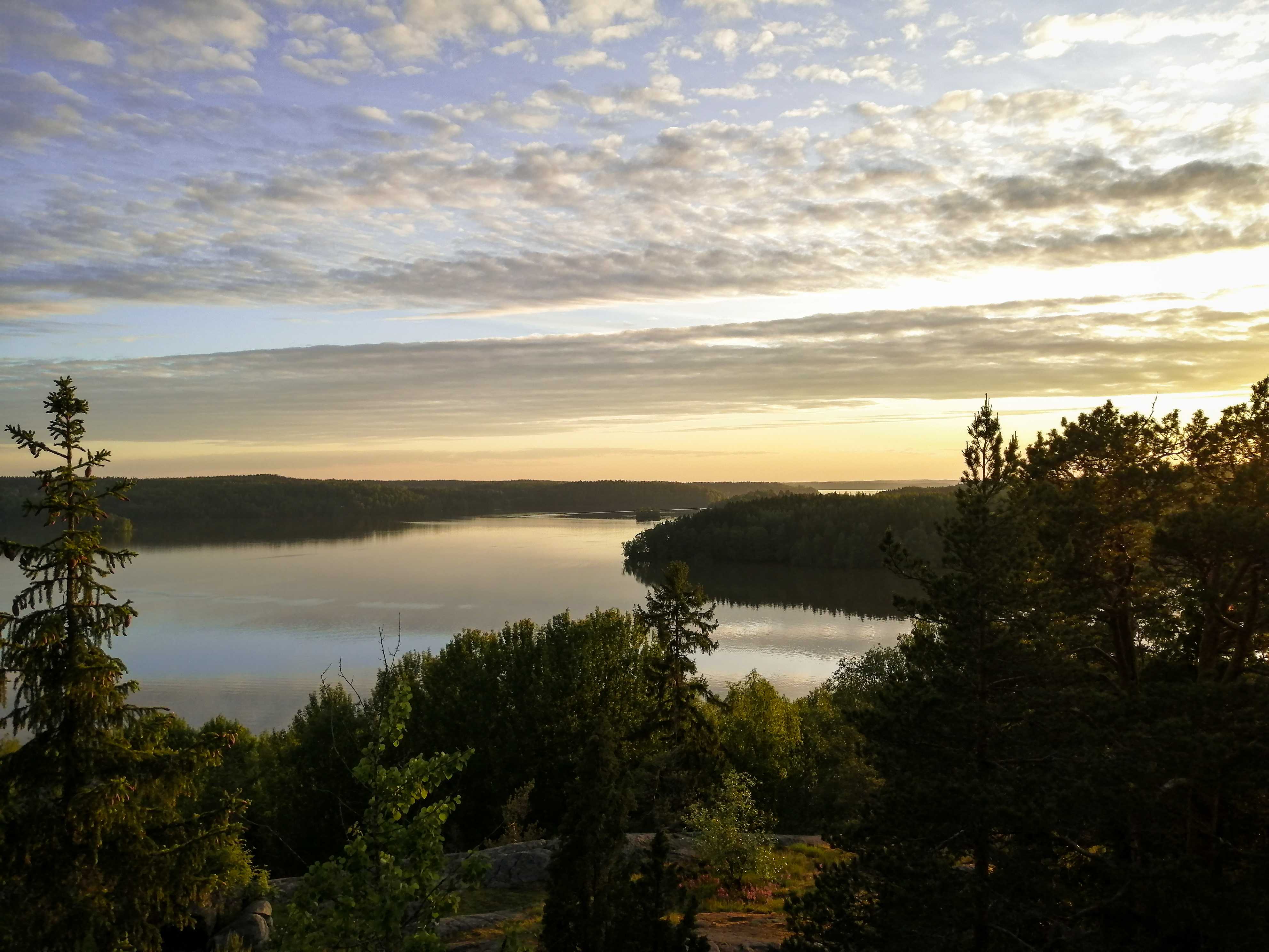 Landscape photo of Hiidenvesi, Finland during sunset. Bottom hald there is a woody downhill leading up to a lake. In the lake there are islands filled with trees. Top half of the image is the sky. The sky gradients from light yellow to blue from bottom to top. The sky is filled with a dotted cloud structure.