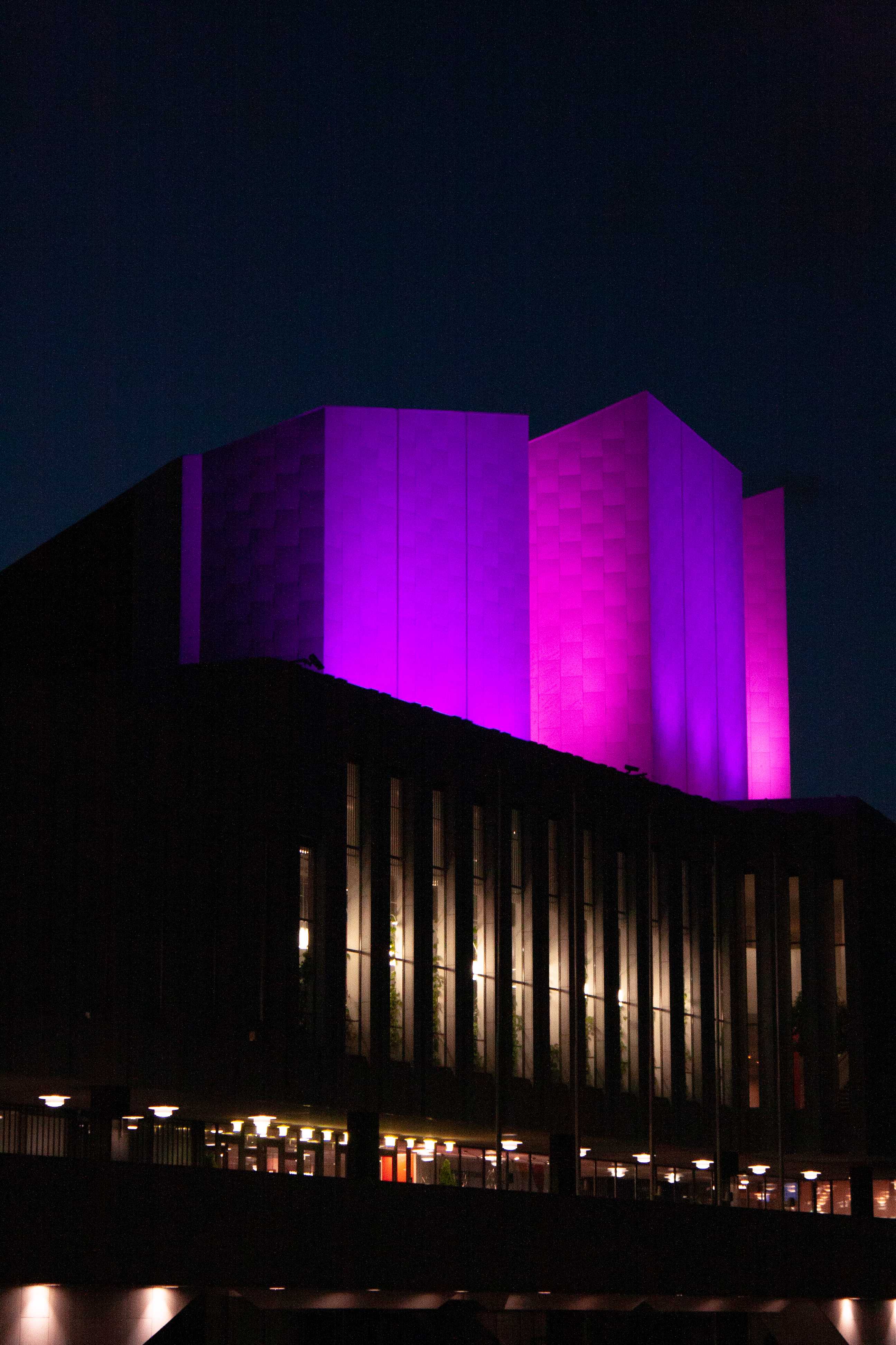 Night picture of the Finlandia building in Helsinki. On the bottom half of the image there is a terrace and above that is a building with tall windows seperated by pillars. On the terrace and inside the hall of which the windows below there are lights. In the top half of the image there is three white, boxy structures lit up in magenta colors. Above that is a dark blue sky.