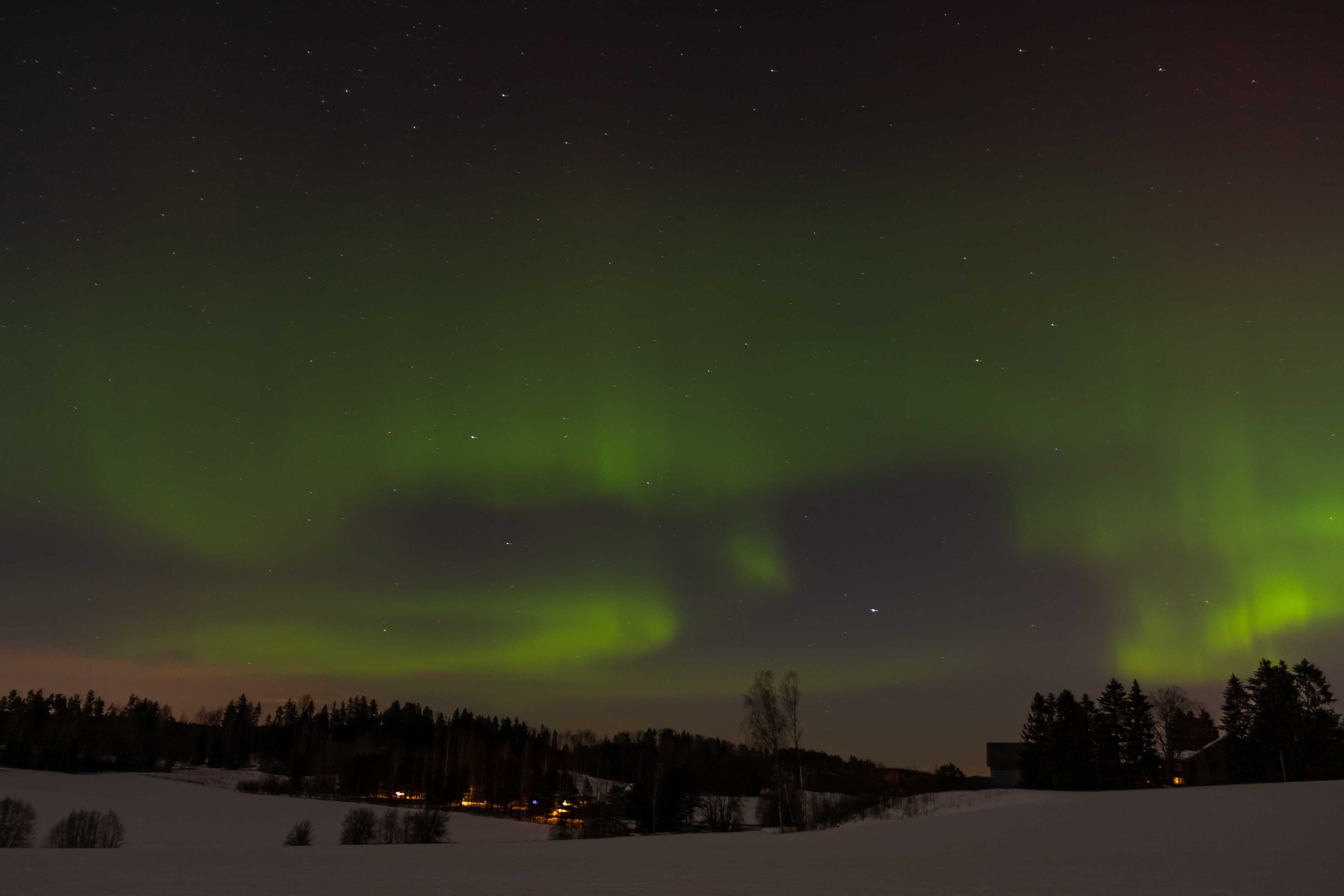 Landscape image of a winter night sky. Sky is filled with aurora borealis which take colors ranging from green to orange to reddish. There are also stars in the sky. On the bottom of the image there is some countryside landscape and couple of yellow lights are visible near the houses in the distance. There is snow on the ground and there is a treeline separating the sky and the ground.